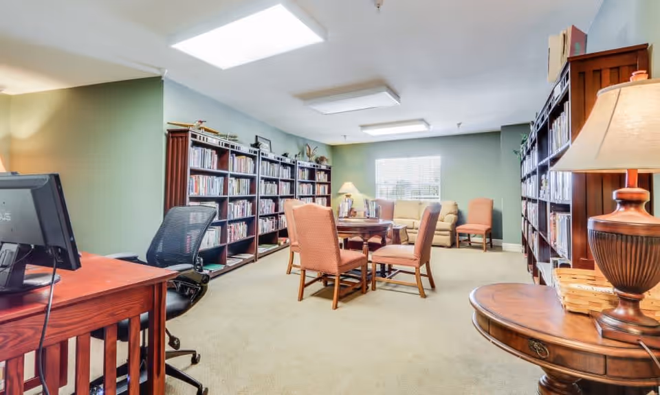 A cozy library or reading room with bookshelves filled with books along the walls, a round wooden table with four upholstered chairs in the center, a beige sofa and an additional chair near a window with blinds, a wooden desk with a computer and office chair, and warm lighting from ceiling fixtures and table lamps.