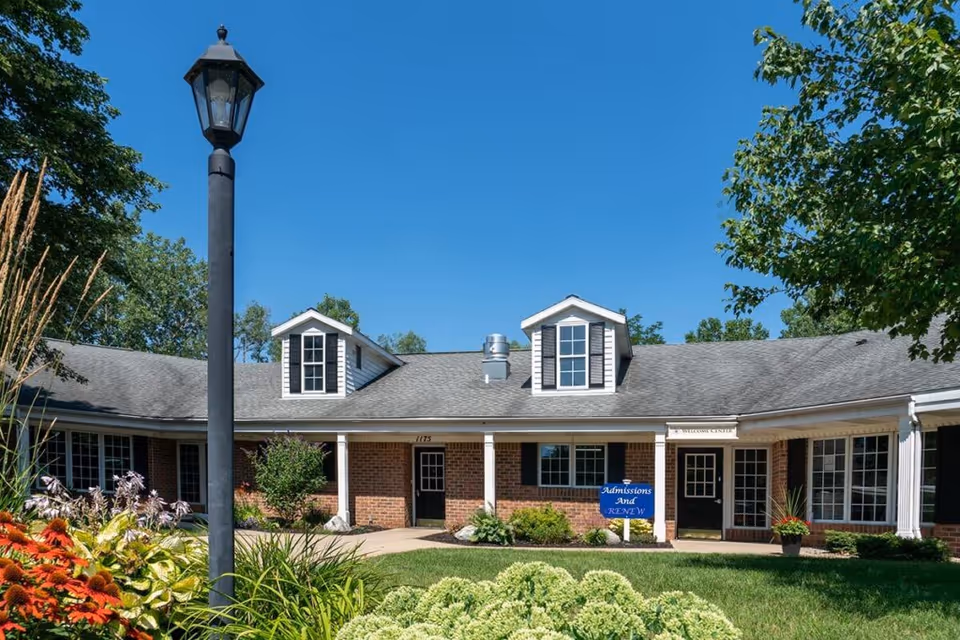 Front exterior of a one-story brick senior living building with dormer windows, landscaped lawn, a lamppost, and a clear blue sky.