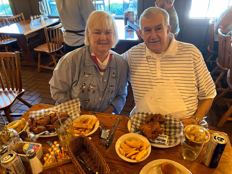 An elderly woman and man sitting together at a wooden table in a restaurant with plates of fried chicken, potato wedges, and drinks in front of them. The woman is wearing a patterned shirt with bird designs, and the man is wearing a white and black striped polo shirt with a napkin tucked in. Other diners and wooden chairs and tables are visible in the background.