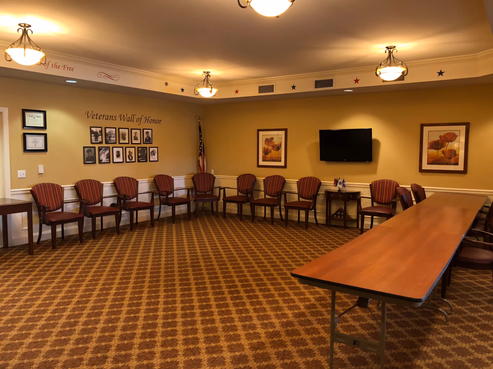 A meeting or activity room with a long wooden table and multiple striped chairs arranged along the walls. The room features a 'Veterans Wall of Honor' with framed photos and an American flag in the corner. There are two framed floral paintings on the wall and a flat-screen TV mounted between them. The ceiling has three light fixtures, and the carpet has a patterned design.