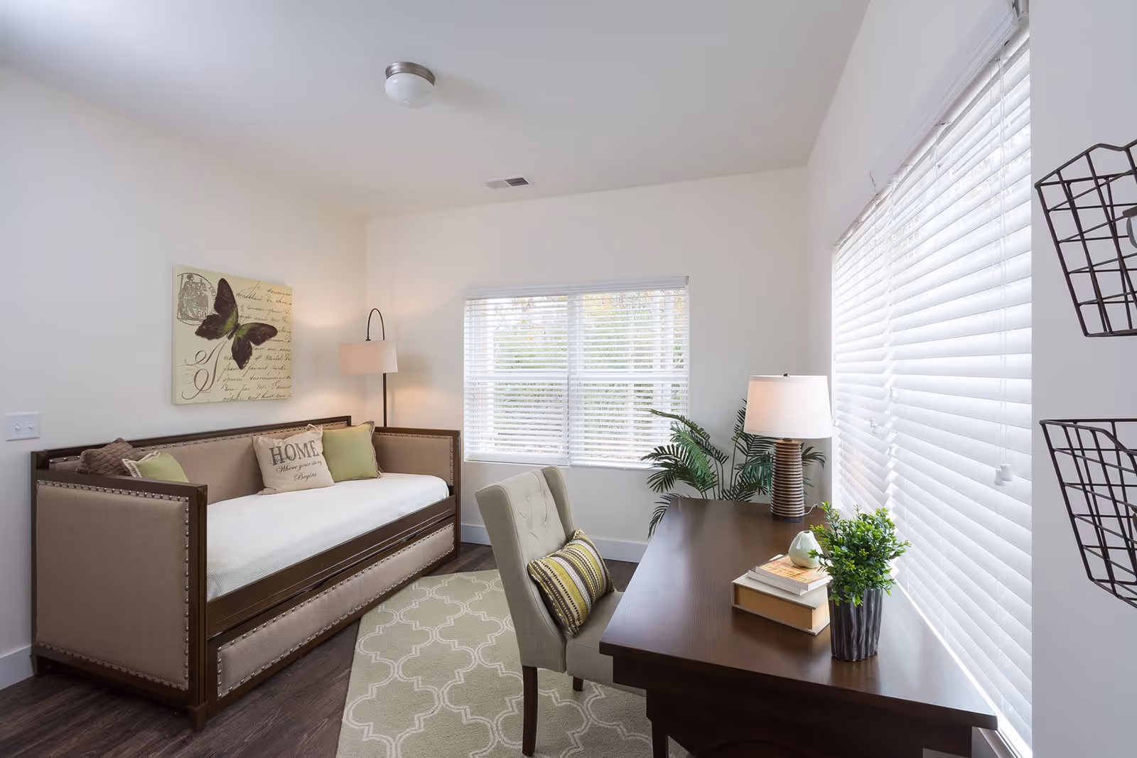A bright room with a daybed featuring decorative pillows, including one with the word 'HOME'. There is a floor lamp beside the bed, a desk with a chair, books, a small plant, and a table lamp. Two windows with white blinds allow natural light to fill the room. The walls are white, and there is a butterfly-themed wall art above the daybed.