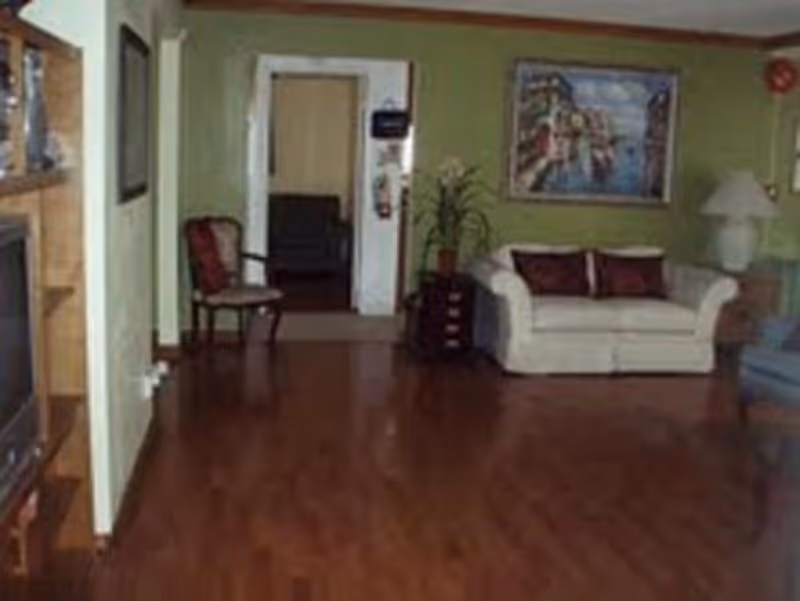 A living room with wooden flooring, a white couch with two dark cushions, a wooden chair with a red cushion, a side table with a lamp, a large framed painting on the green wall, and a television on a wooden stand.