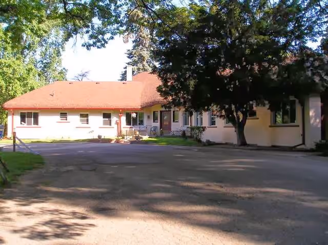 Exterior view of a single-story building with a red roof and cream-colored walls, surrounded by trees and greenery, with a paved driveway in front.