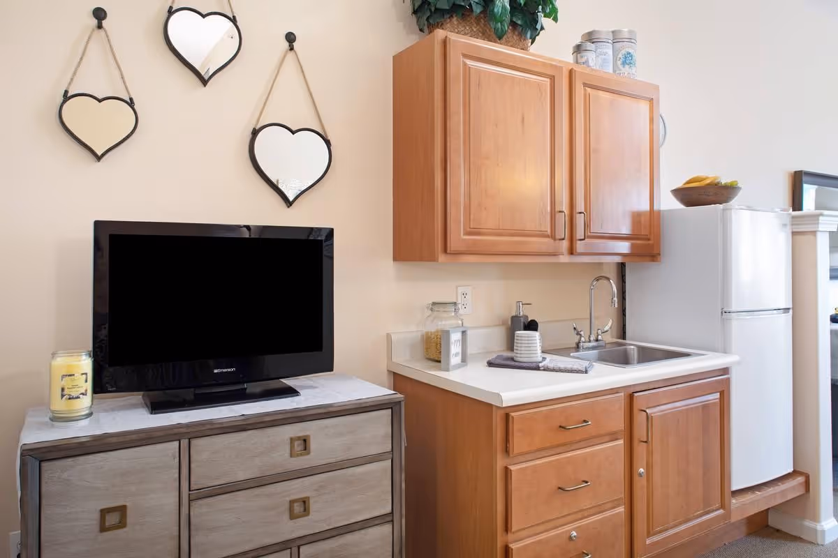 A small kitchenette area with wooden cabinets, a white countertop, a sink, and a white refrigerator. Next to the kitchenette is a wooden dresser with a flat-screen TV on top. Three heart-shaped mirrors hang on the wall above the TV. A candle and some decorative items are also visible on the dresser and countertop.