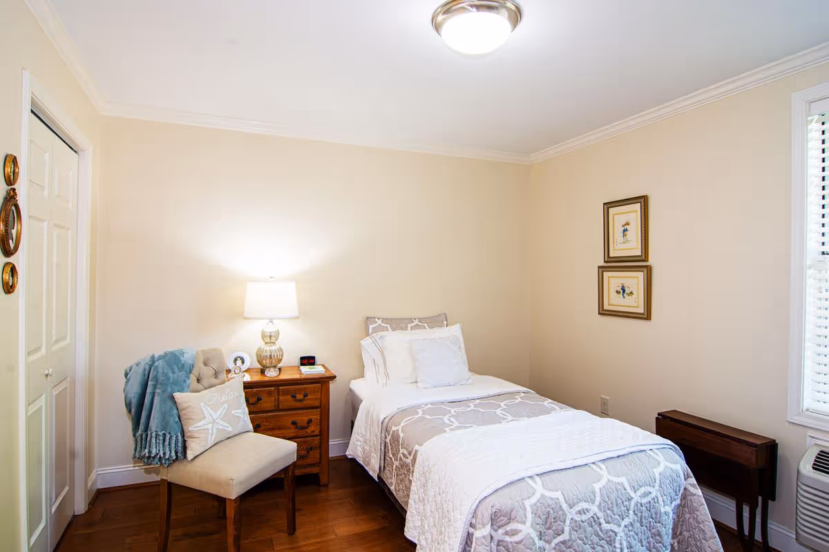 Well-lit single bedroom with a twin bed, wooden nightstand and lamp, an upholstered chair with a throw, and framed wall art.