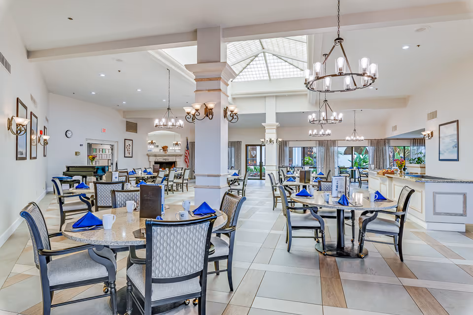 Bright, spacious dining room with multiple round tables set with blue napkins and chandeliers in a senior living facility.
