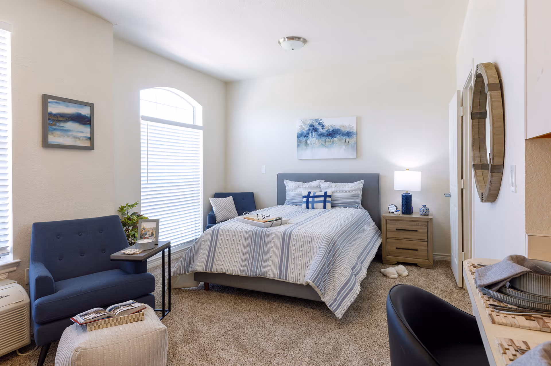 A bright and cozy bedroom featuring a bed with striped bedding and multiple pillows, a wooden nightstand with a lamp and decorative items, a blue armchair with a small side table, a white ottoman with an open book, a large window with blinds, a round wall mirror, and a piece of abstract artwork above the bed.