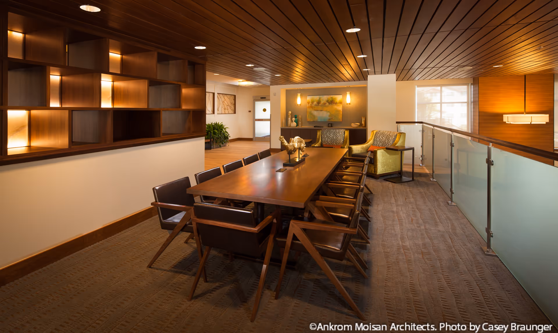 A modern, well-lit interior room with a long wooden table surrounded by twelve dark brown chairs. The room features a wooden ceiling with recessed lighting, a decorative wooden shelving unit on the left wall, and two green armchairs with patterned cushions near the back wall. There is a glass railing on the right side and artwork on the walls, creating a warm and inviting atmosphere.