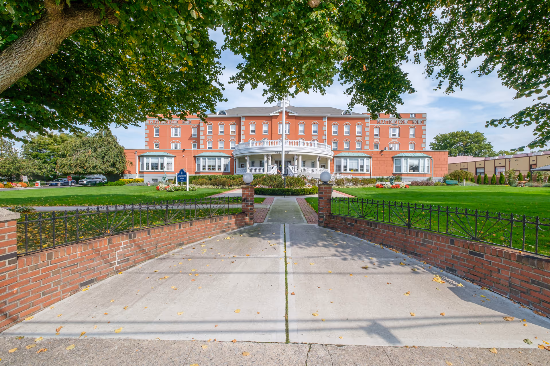 Front view of a large red-brick senior living building with a circular porch, driveway, and manicured lawn framed by trees.