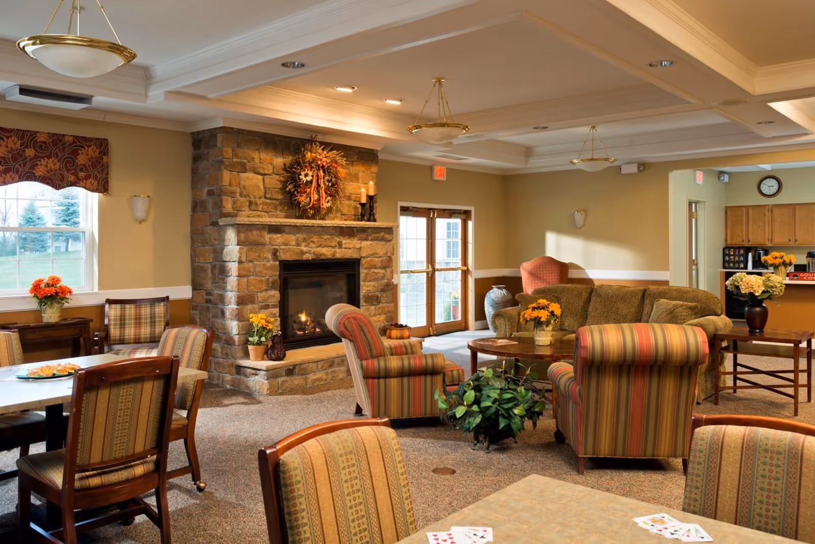 A cozy living room area in a senior living facility with a stone fireplace decorated with a wreath and candles. The room features multiple striped upholstered armchairs, a green sofa, wooden tables with flower arrangements, and a window with a patterned valance. The space is warmly lit with ceiling lights and has a carpeted floor. In the background, there is a kitchenette area with cabinets and a clock on the wall.