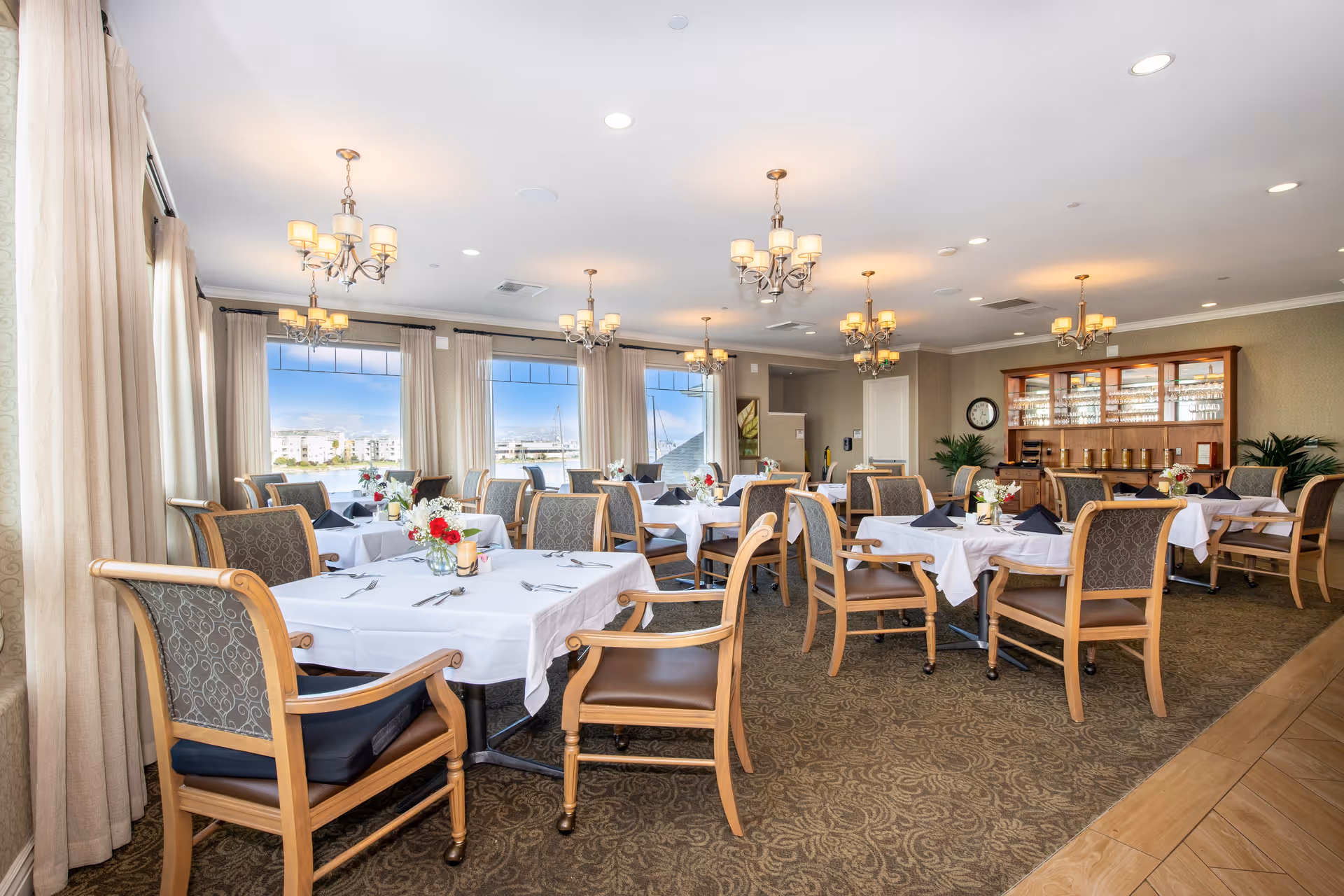 A bright and elegant dining room with multiple tables covered in white tablecloths, each set with silverware, napkins, and small floral centerpieces. The room features large windows with sheer curtains allowing natural light to fill the space, chandeliers hanging from the ceiling, and a wooden cabinet with glassware and coffee dispensers against the far wall.