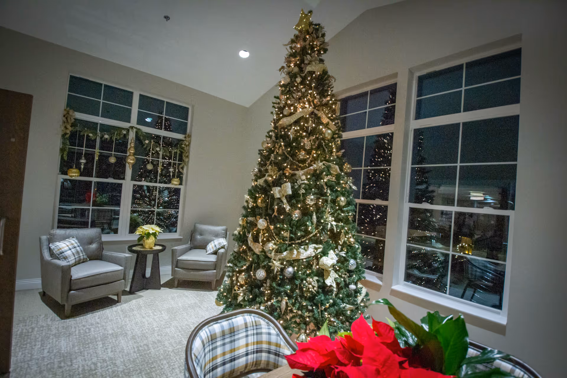 A cozy room decorated for Christmas with a tall, decorated Christmas tree adorned with lights, ribbons, and ornaments. Two gray armchairs with plaid pillows sit near a window with holiday garland. A table with a red poinsettia plant is partially visible in the foreground.