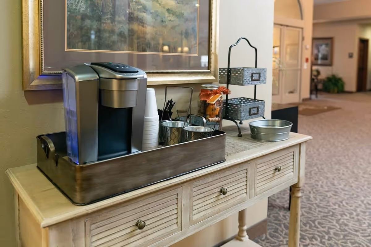 A coffee station on a wooden table with a coffee maker, disposable cups, stirrers, and metal containers. The table is placed in a hallway with carpeted flooring and a framed painting on the wall above the table.