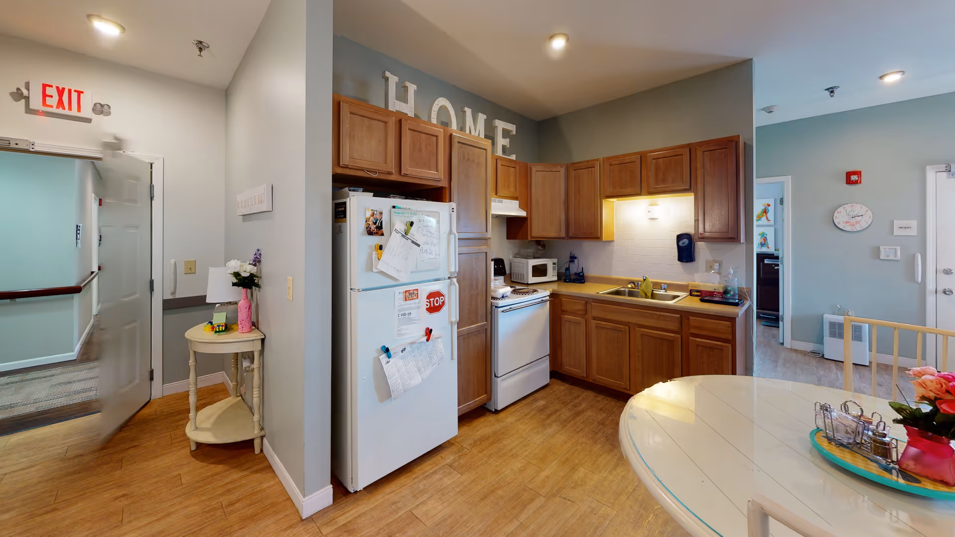Interior view of a senior living facility kitchen area with wooden cabinets, a white refrigerator covered with papers and magnets, a stove, microwave, and a sink. A round dining table with a flower vase and condiments is partially visible in the foreground. To the left, there is an open door leading to a hallway with an exit sign above it. The walls are painted light gray and the floor is wooden. The word 'HOME' is displayed on top of the cabinets.