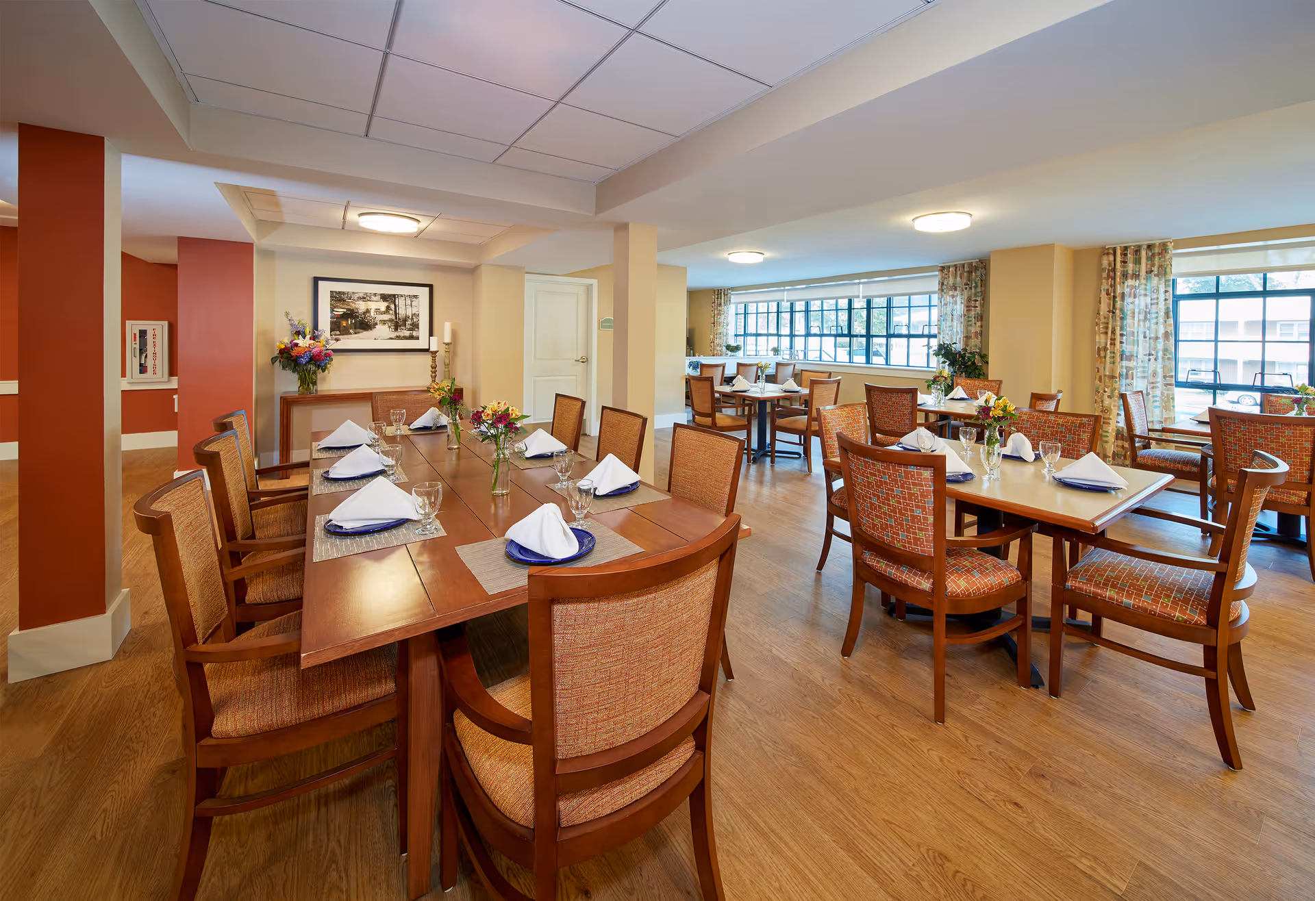 A bright and spacious dining room in a senior living facility with multiple wooden tables and chairs. Each table is set with white folded napkins, glasses, and small floral centerpieces. Large windows with patterned curtains allow natural light to fill the room, and the floor is made of wood. The walls are painted in warm tones, and there is a framed black and white photograph and a flower arrangement on a side table against the wall.