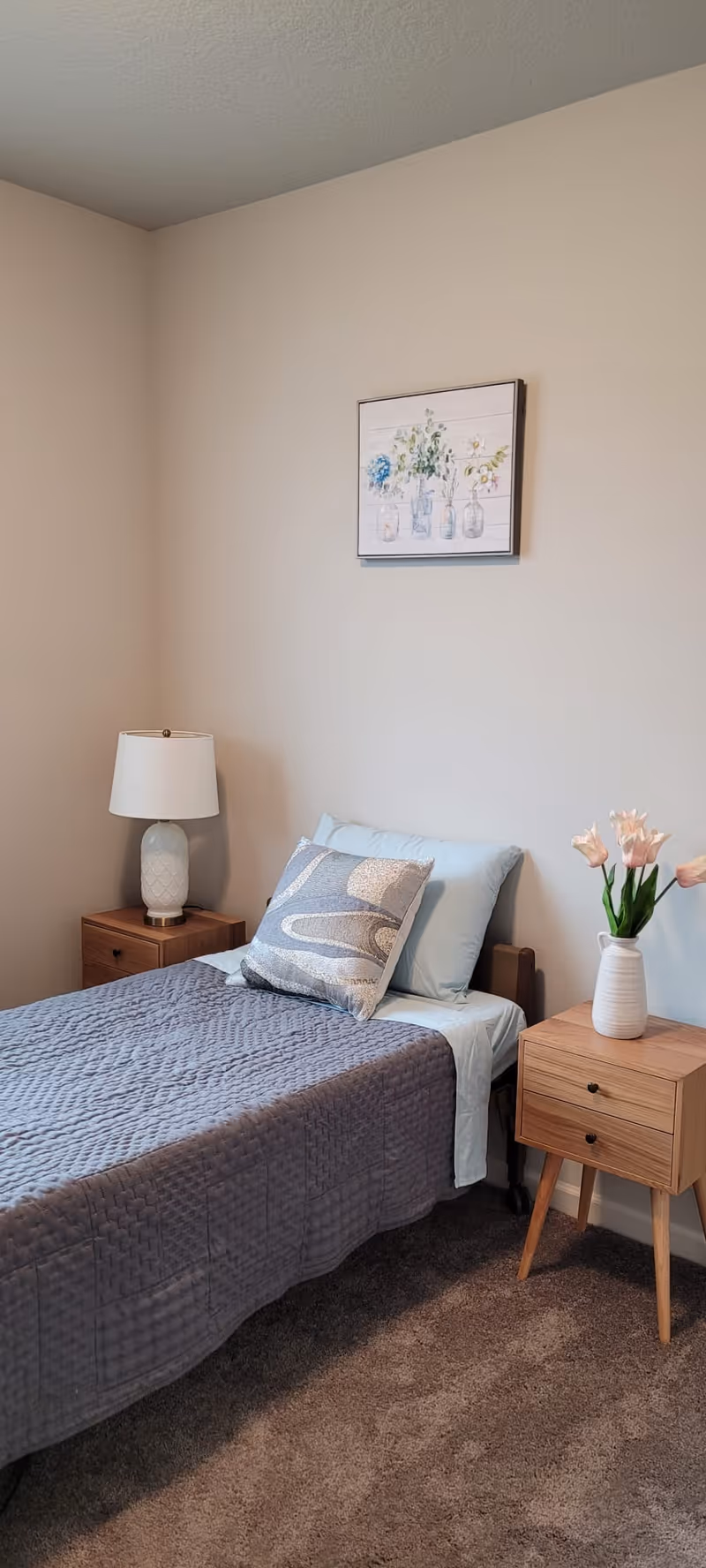 A neatly made single bed with a gray quilted bedspread and two pillows, one light blue and one patterned. On the left side of the bed is a wooden nightstand with a white lamp. On the right side is a small wooden side table with a white vase holding pink flowers. Above the bed hangs a framed picture of flowers in jars. The room has beige walls and carpeted floor.