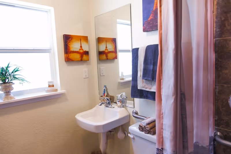 A small bathroom with a white sink, a mirror above it, a window with a potted plant on the sill, a towel rack with blue and white towels, a toilet with folded towels on top, and a pink shower curtain partially visible.