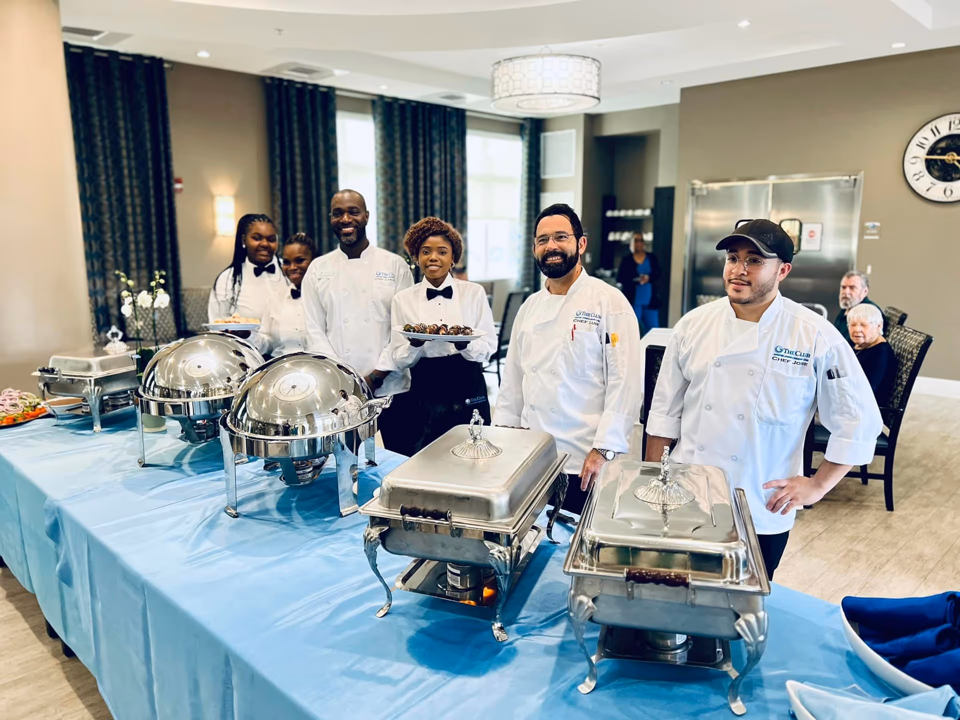Catering staff and chefs stand behind a buffet table with chafing dishes in a dining room.