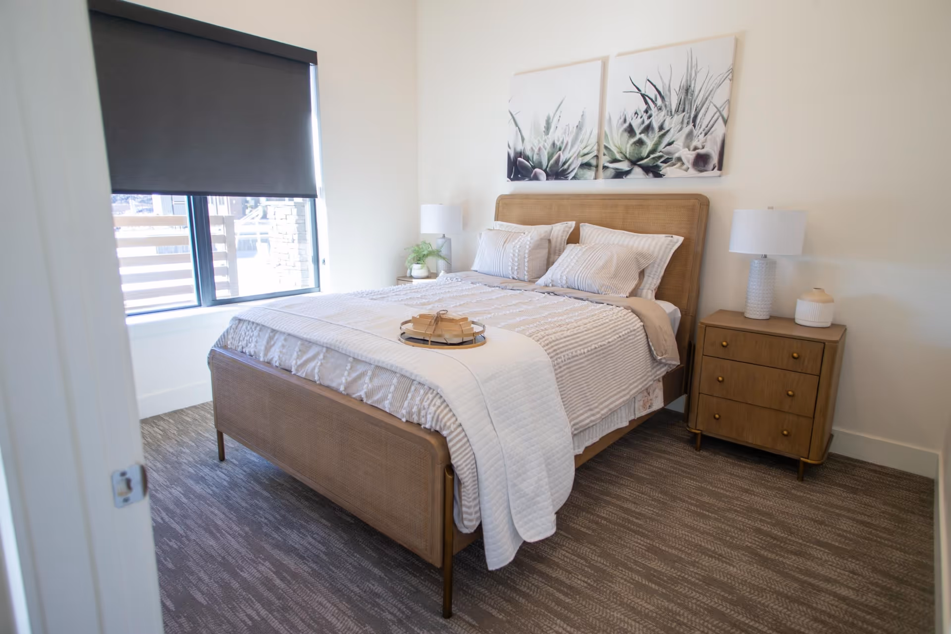 A neatly made bed with beige and white bedding in a bedroom. The bed has a wooden frame and is flanked by a wooden nightstand with a white lamp and decorative items. Above the bed are two framed botanical prints. A window with a dark roller shade is on the left side of the room.