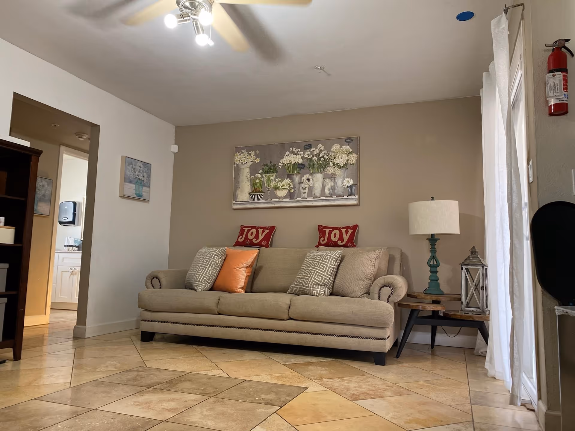Cozy living room with a beige sofa decorated with throw pillows that say 'JOY', a side table and lamp, wall art, and tiled tile floor.