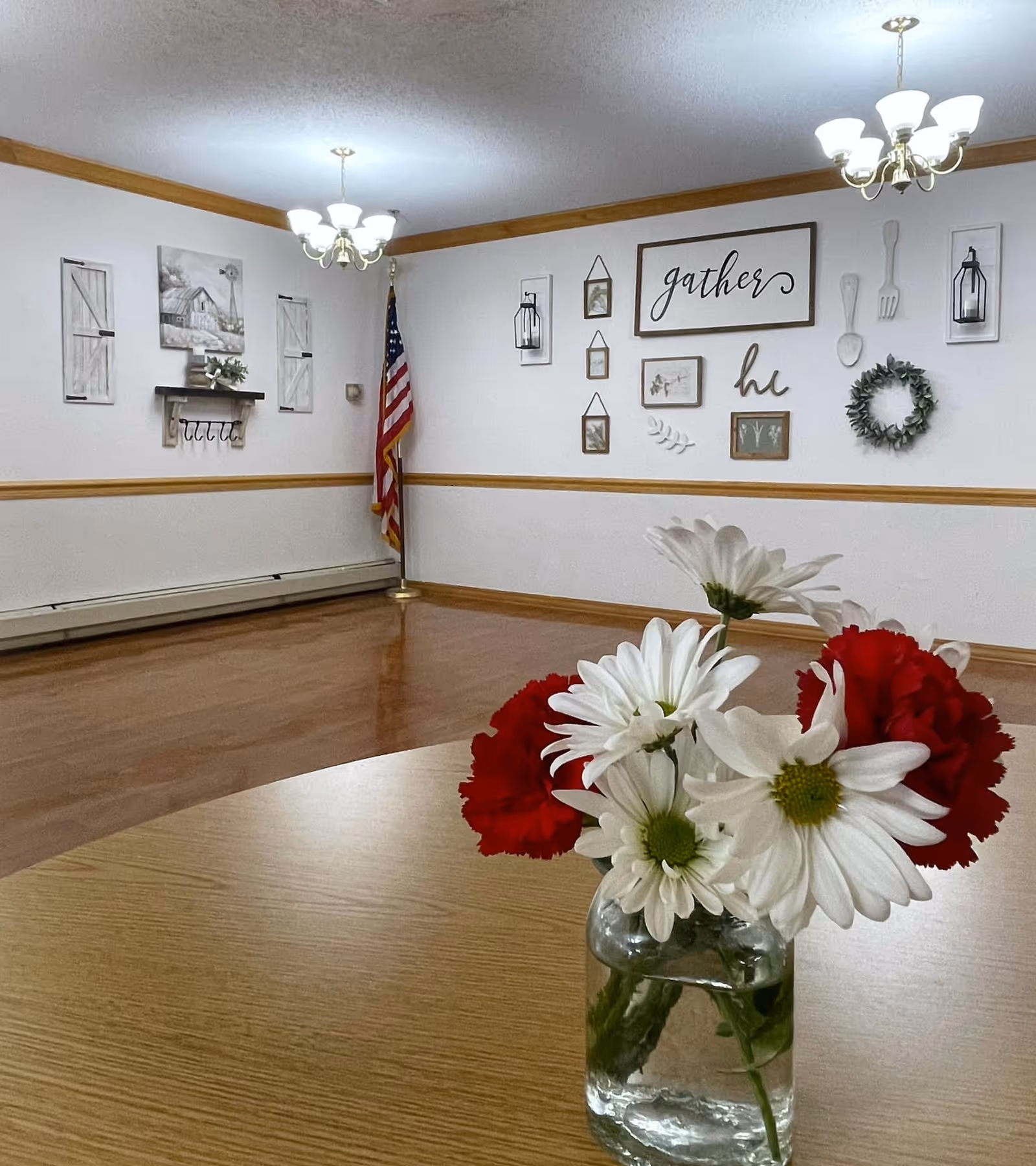 A round wooden table with a small glass vase holding white daisies and red carnations in the foreground. The background shows a room with light-colored walls, wooden trim, two ceiling light fixtures, an American flag in the corner, and wall decorations including framed pictures, a wreath, and a sign that says 'gather'.