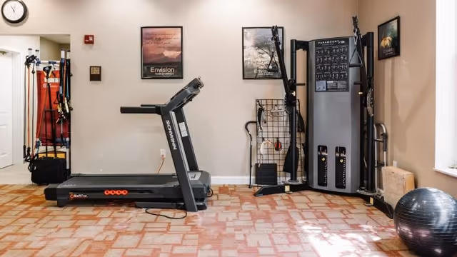 A fitness room with a treadmill, a multi-function weight machine, exercise bands hanging on a rack, and a large exercise ball on a patterned carpeted floor. The walls have framed motivational posters and a clock above a doorway.