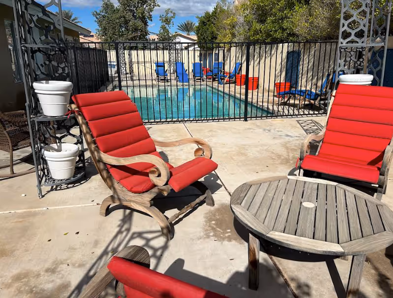 Outdoor pool area with red-cushioned wooden lounge chairs and a round wooden table by a fenced swimming pool.