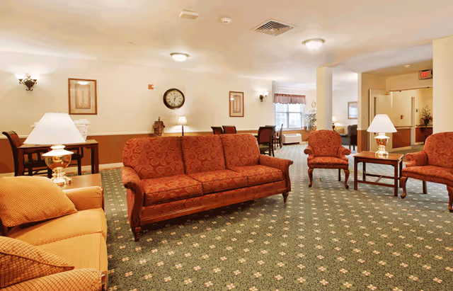 A spacious senior living lounge with patterned sofas, armchairs, side tables, lamps, and a clock on the wall.