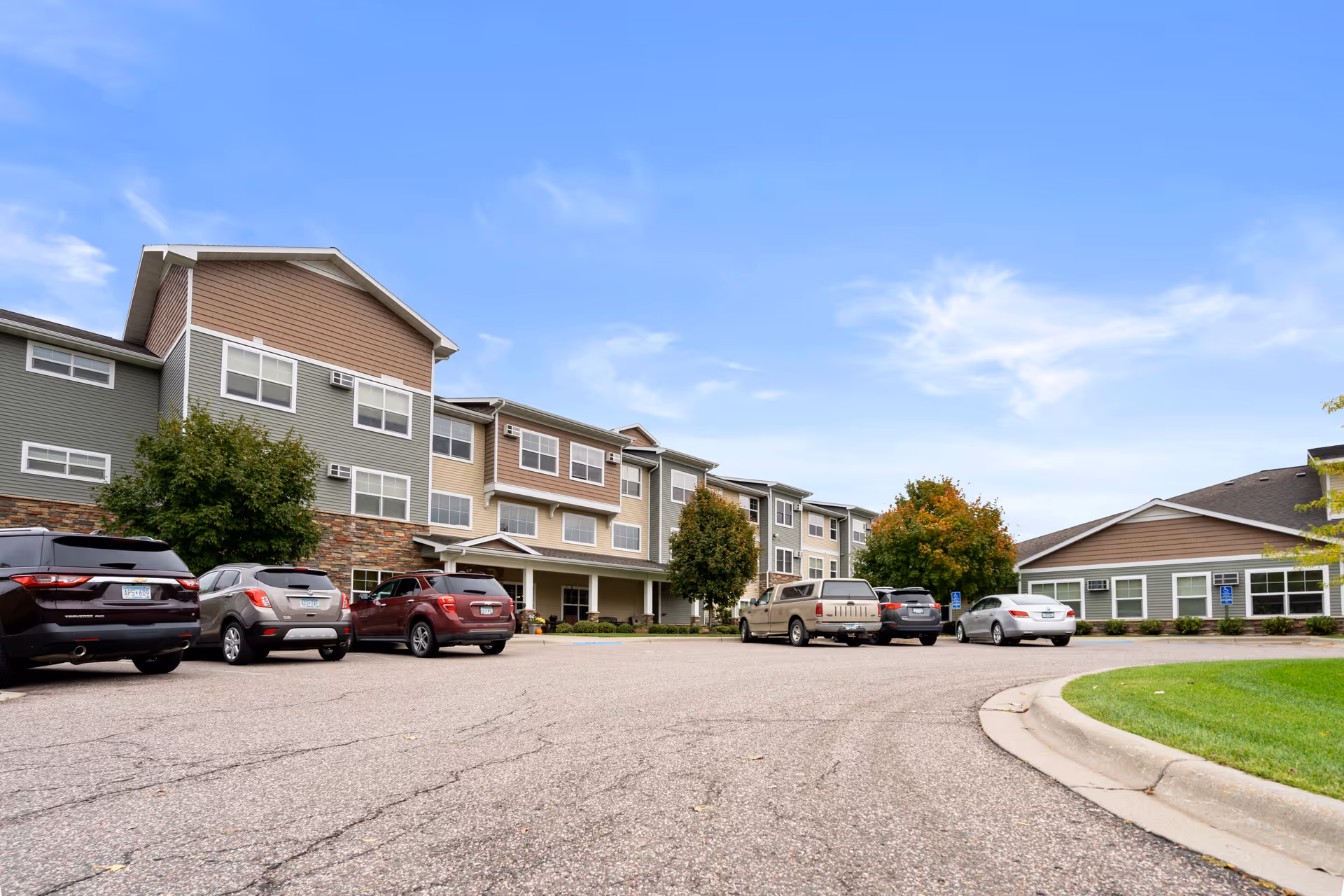 Exterior view of a senior living facility named Legacy of Delano Senior Living, showing a multi-story building with beige and green siding, several parked cars in the parking lot, trees with some autumn foliage, and a clear blue sky.