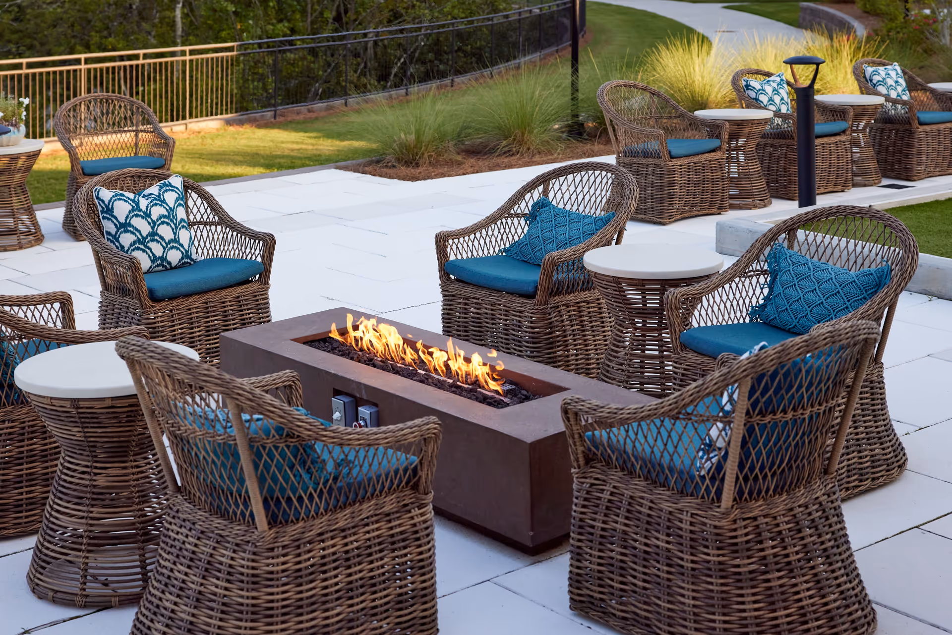 Outdoor patio with wicker chairs and blue cushions arranged around a rectangular fire pit on a tiled terrace