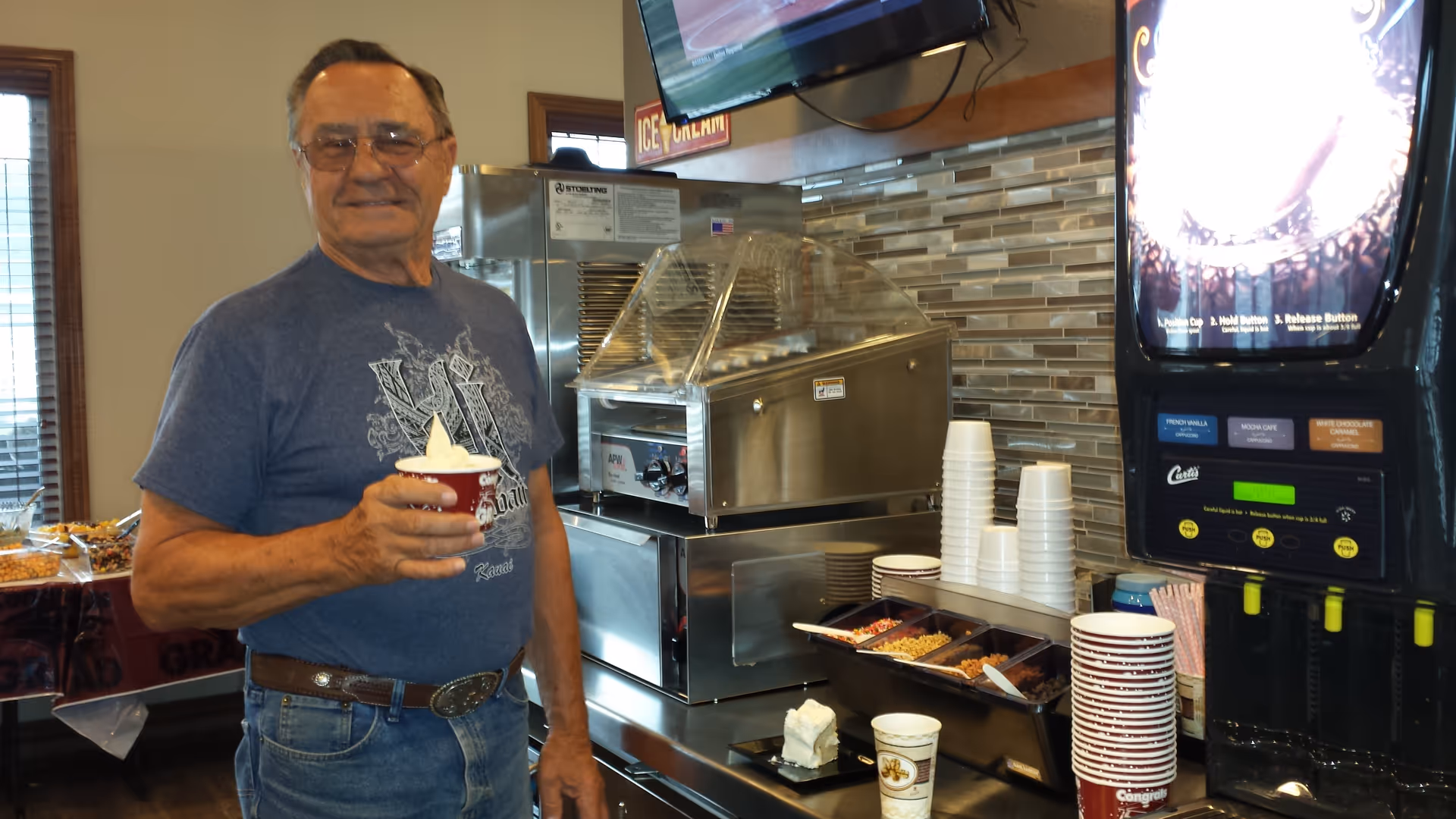 An elderly man wearing glasses and a blue t-shirt stands indoors holding a cup of soft serve ice cream. Behind him is a counter with a soft serve ice cream machine, a coffee machine, stacks of cups, and various toppings. The setting appears to be a casual dining or snack area.
