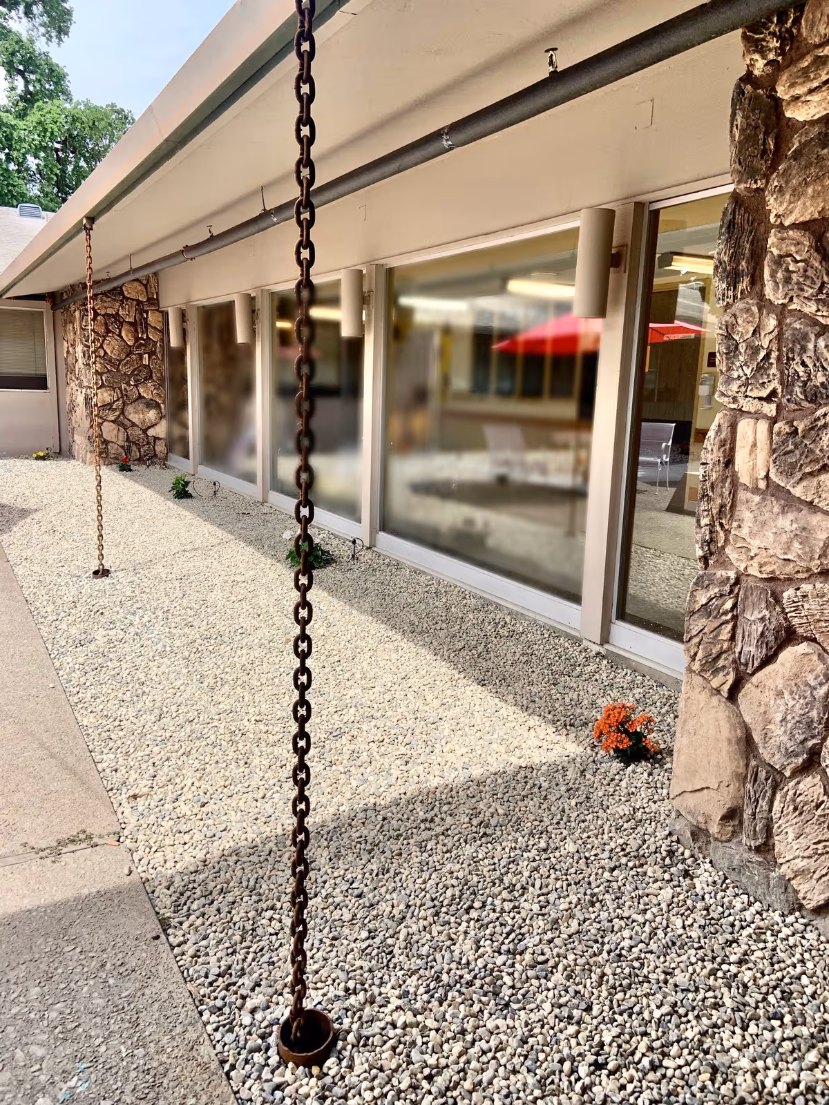 Outdoor area of a facility with a gravel ground, stone wall, large windows, and two hanging metal chains attached to the roof. There are some small plants near the windows and a red umbrella reflected in the glass.