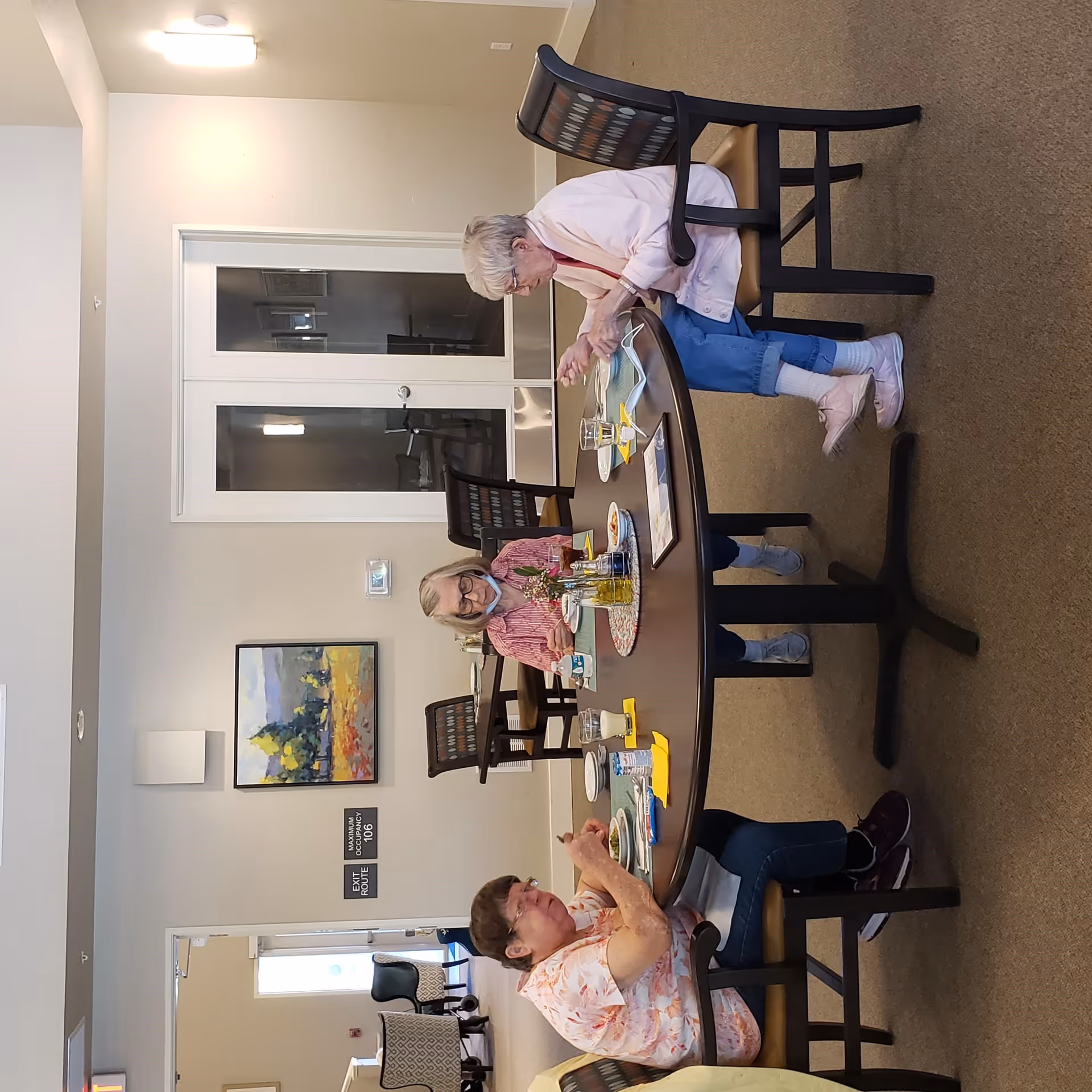Three elderly women sitting around a dark wooden dining table in a well-lit room, eating and conversing. The room has beige walls, a carpeted floor, and a painting on the wall. There are additional chairs and a door in the background.