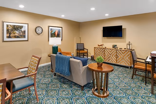 A cozy common lounge with sofas and chairs arranged around side tables, a wall-mounted TV, decorative artwork, and patterned carpet.