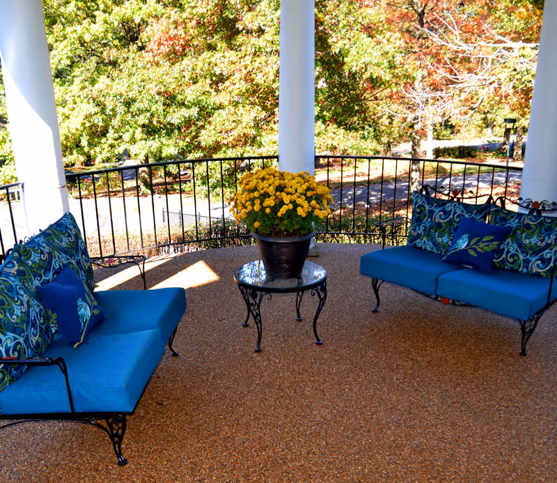 Outdoor covered patio area with two blue cushioned wrought iron benches facing each other, a small round glass-top table with a pot of yellow flowers in the center, surrounded by black metal railing and trees with autumn foliage in the background.