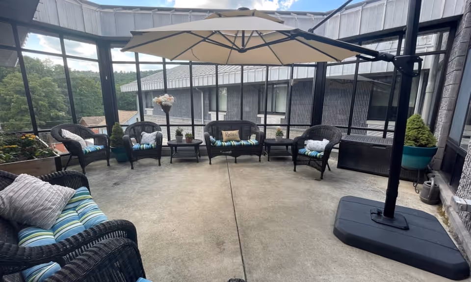 A screened-in outdoor patio area with several black wicker chairs and a loveseat, all with blue and green striped cushions and decorative pillows. A large beige umbrella provides shade over the seating area. There are potted plants and flowers around the perimeter, and a building with windows is visible in the background.