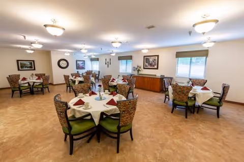 A spacious dining room with multiple round tables covered with white tablecloths and set with cups, plates, and folded red napkins. Each table is surrounded by patterned chairs with green cushions. The room has beige walls, several windows with blinds, framed artwork, and ceiling lights providing bright illumination.