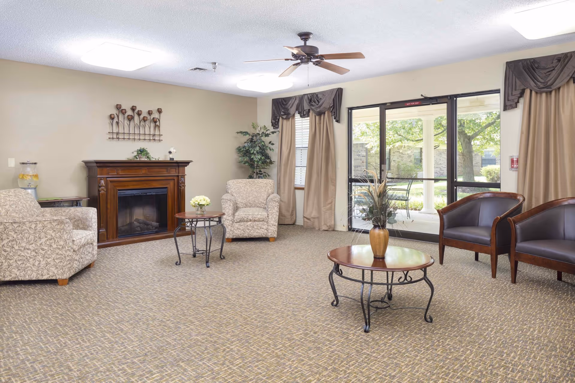 A senior living facility common area with beige walls and carpeted floor. The room features a wooden fireplace, two patterned armchairs, two dark brown leather chairs, two round wooden tables with metal legs, a ceiling fan, and large windows with beige curtains overlooking an outdoor patio with trees and chairs.