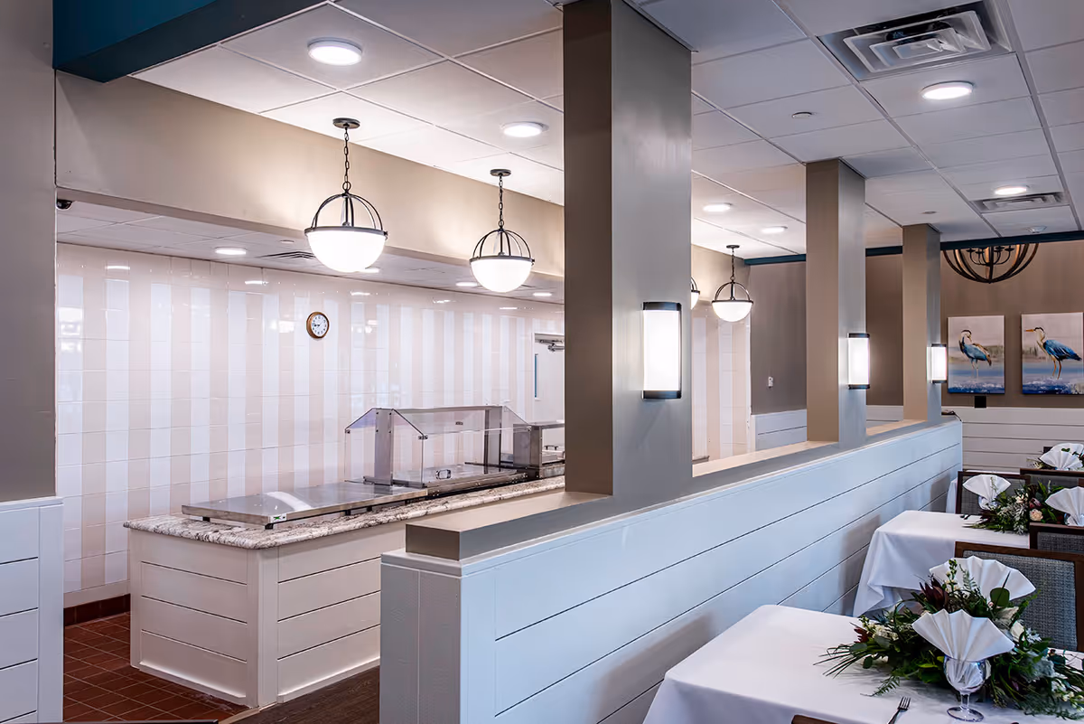 Dining room with a serving counter, pendant lights, and tables set with folded napkins and floral centerpieces.