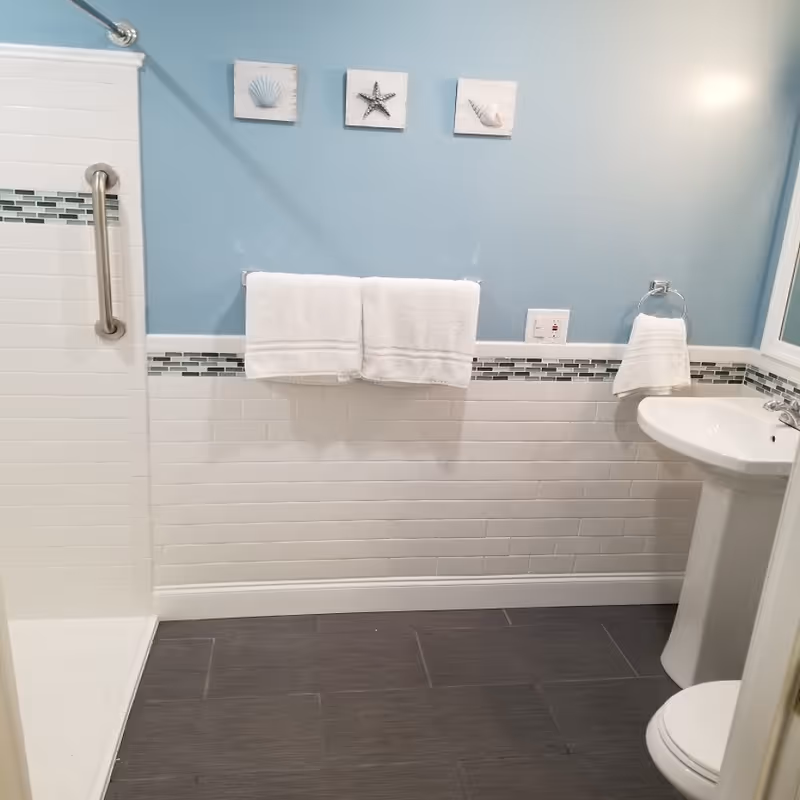A clean and modern bathroom with light blue walls and white subway tile halfway up the wall. There is a walk-in shower with a grab bar, two white towels hanging on a towel rack, a white pedestal sink with a hand towel on a ring, and a toilet partially visible. Three small framed pictures of seashells and a starfish are hung on the wall above the towels.