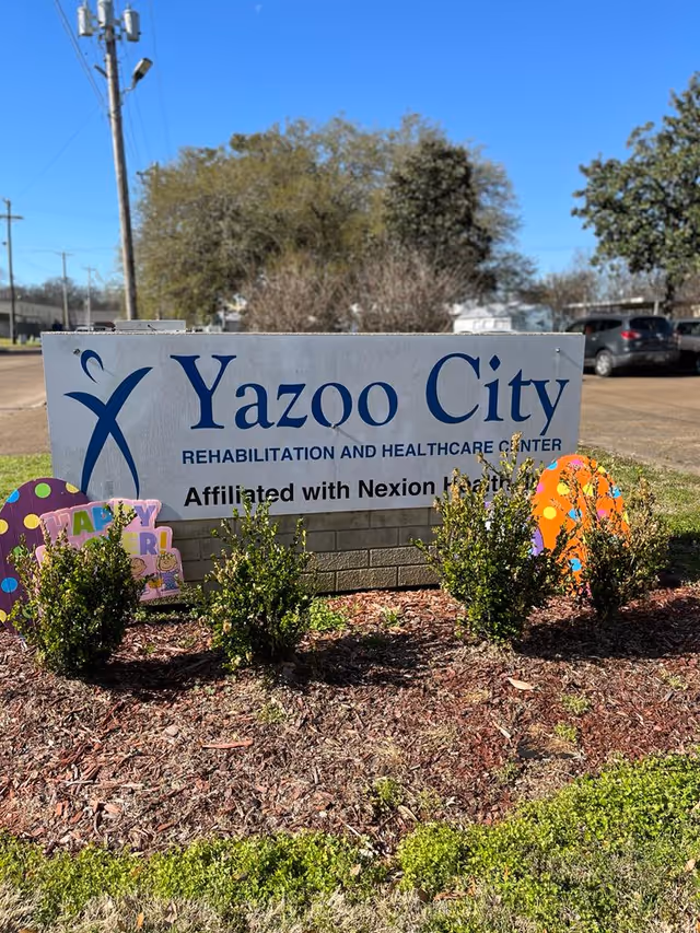 Outdoor view of a sign for Yazoo City Rehabilitation and Healthcare Center, affiliated with Nexion Health, surrounded by small bushes and decorative signs on a sunny day with a clear blue sky.