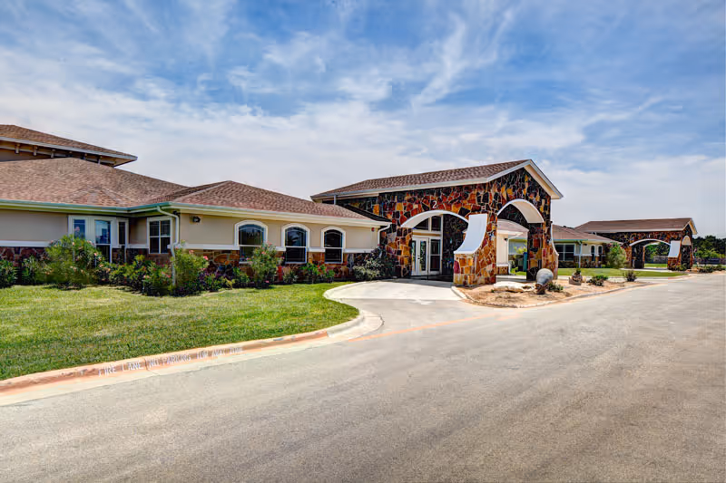 Exterior view of a single-story assisted living facility with a stone and stucco facade, arched entryways, a driveway, and well-maintained landscaping under a partly cloudy sky.