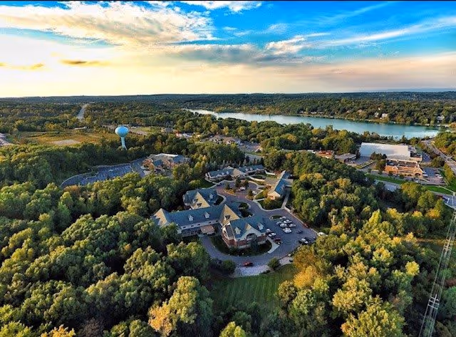 Aerial view of a senior living facility surrounded by dense green trees with a river visible in the background under a partly cloudy sky during daytime.