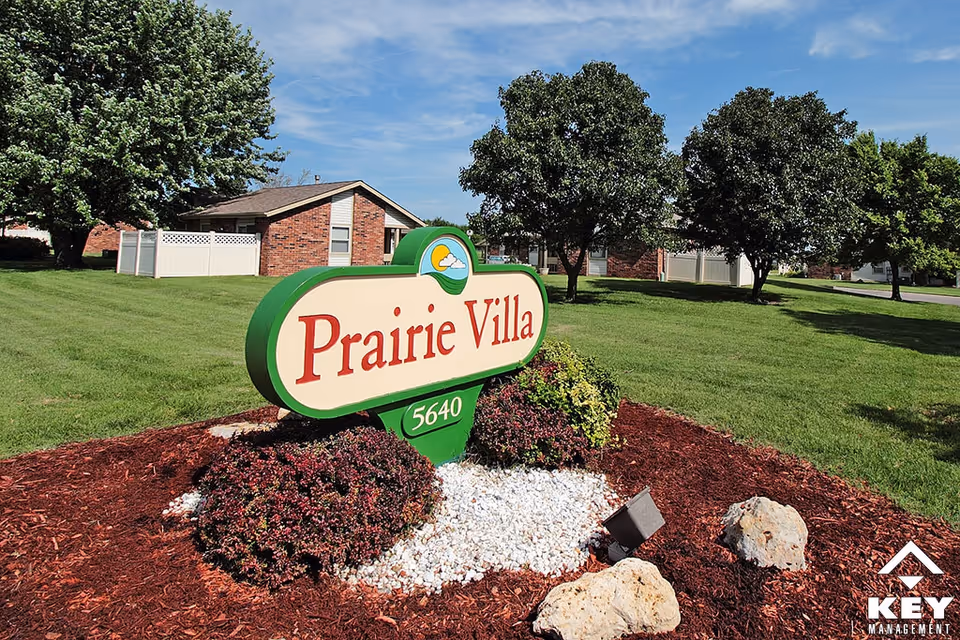 Outdoor view of the Prairie Villa senior living facility sign surrounded by landscaping with bushes, rocks, and mulch. In the background, there are green lawns, trees, and brick buildings under a blue sky with some clouds.