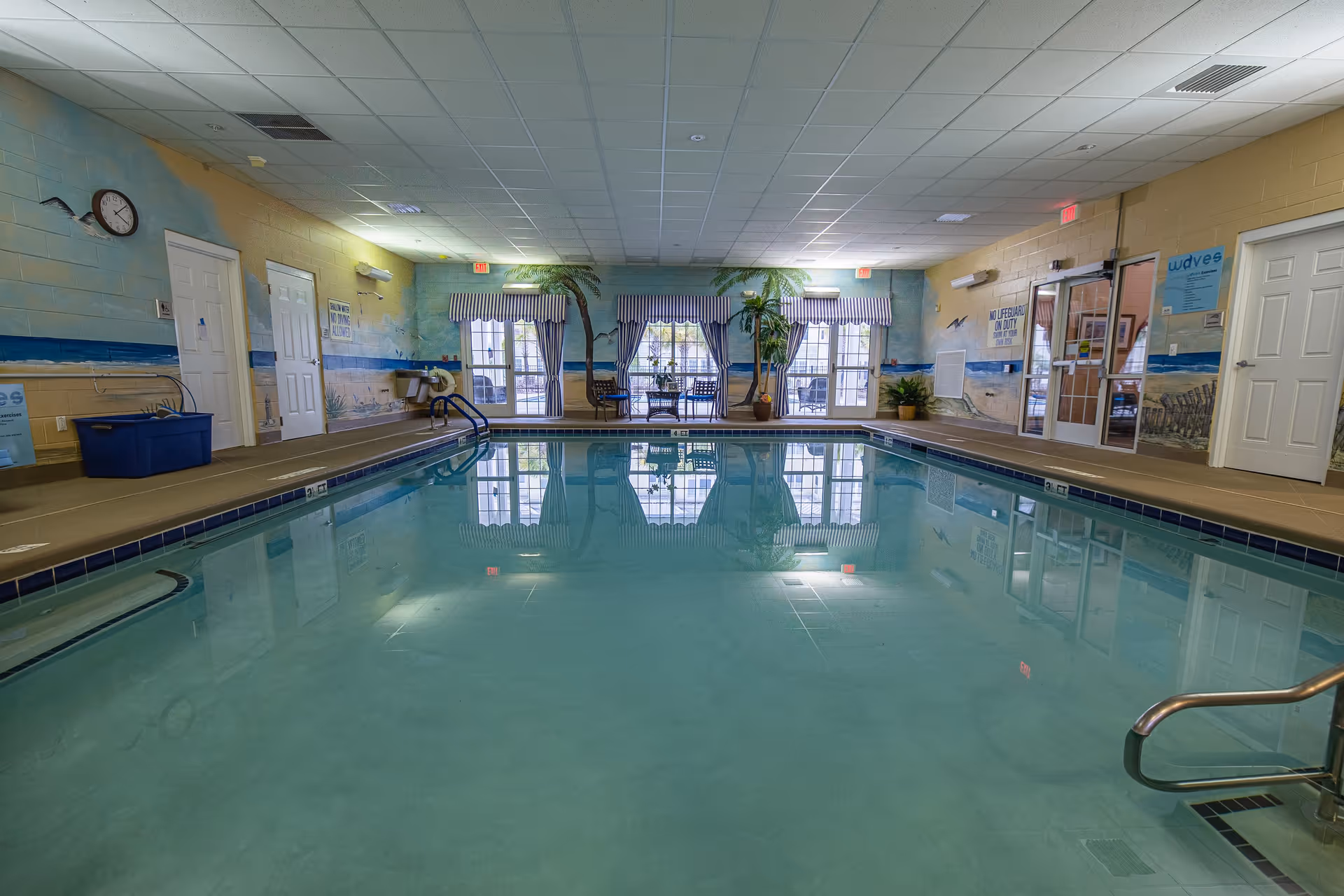 Indoor swimming pool area with clear water, surrounded by a beige tiled floor and walls painted with a beach and ocean mural. There are several doors and windows with striped blue and white curtains, and some potted plants and chairs near the windows. The ceiling has white tiles with recessed lighting.