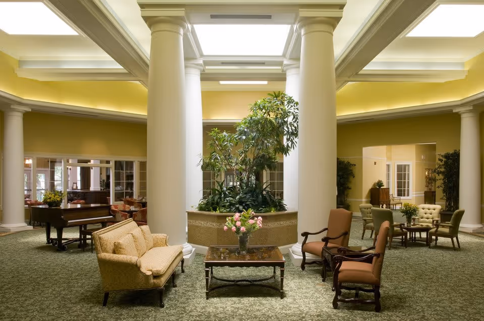 A spacious and elegant common area with large white columns surrounding a central planter filled with green plants. The room features a green patterned carpet, beige and brown upholstered chairs and sofas arranged around wooden coffee tables, and a grand piano in the background. The walls are painted yellow with white trim, and large windows and doorways lead to other rooms.