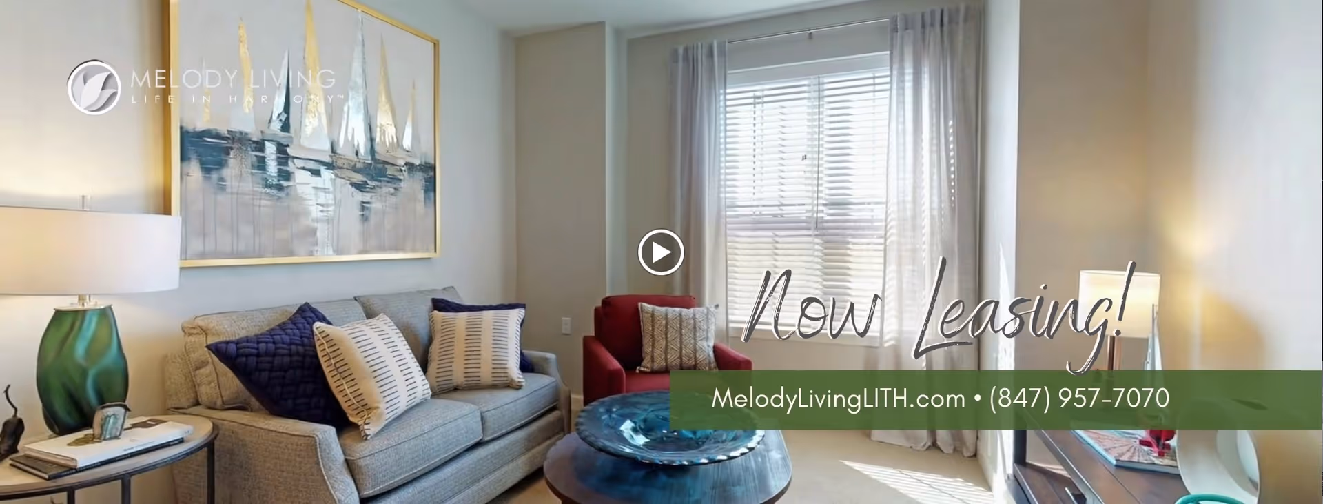 A cozy living room with a beige sofa adorned with multiple pillows, a green table lamp on a side table, a red armchair, a round coffee table, and a large window with white blinds and sheer curtains allowing natural light to fill the room. A large painting of sailboats hangs on the wall above the sofa.