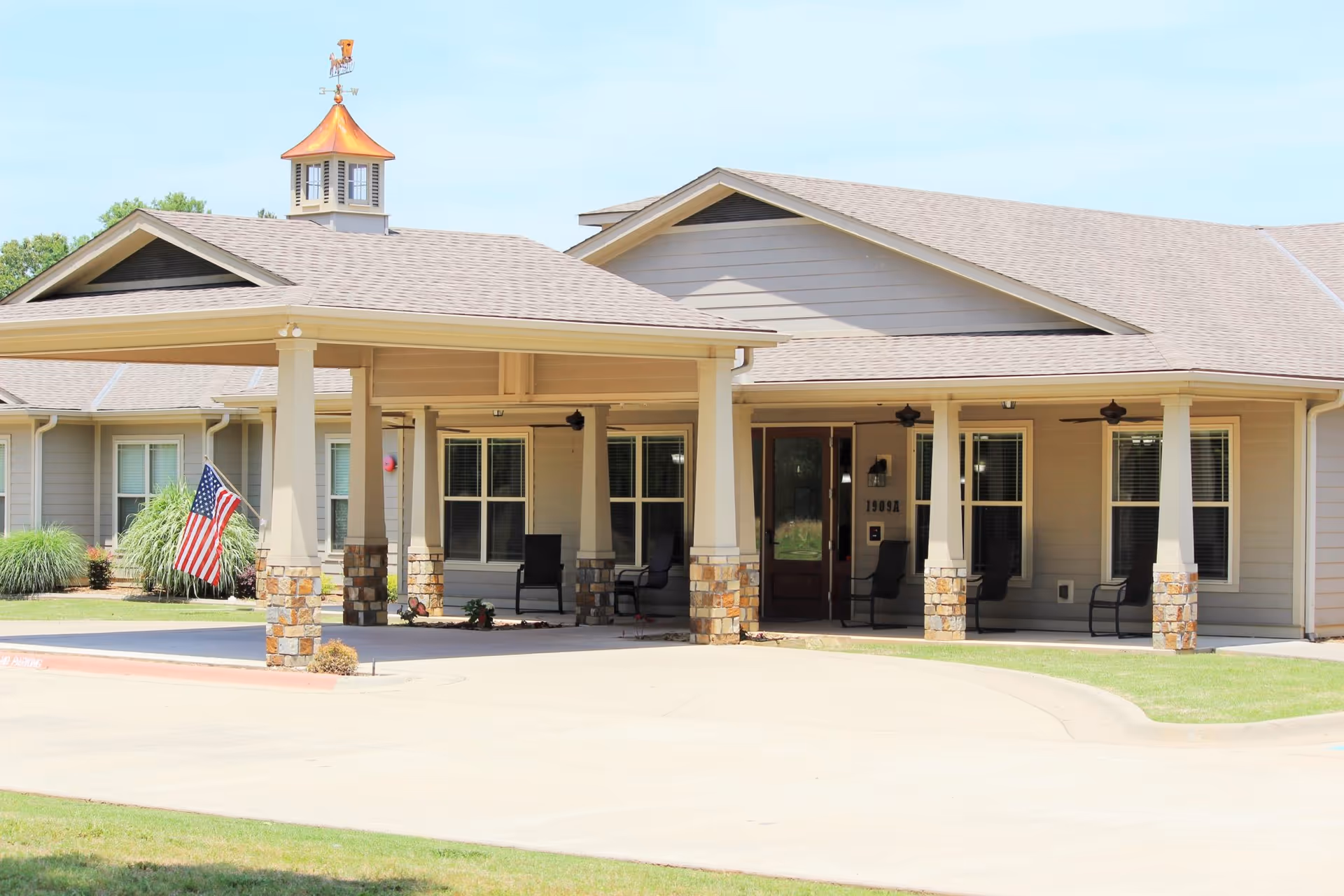 Front entrance of a single-story senior living building with a covered porte-cochere, stone-columned porch, American flag, and outdoor seating.