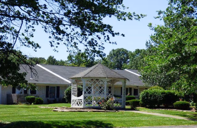 A white gazebo on a manicured lawn in front of a single-story retirement village building surrounded by trees.