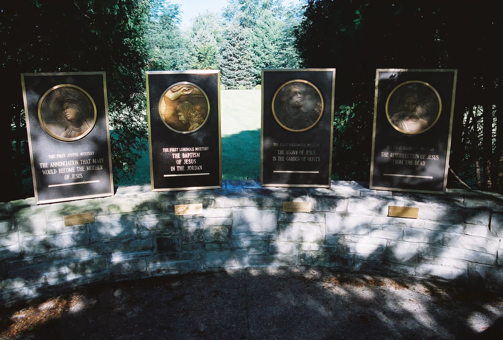 Four bronze plaques mounted on a curved stone wall outdoors, each depicting a religious scene related to Jesus with inscriptions describing different mysteries: The Annunciation that Mary would become the mother of Jesus, The Baptism of Jesus in the Jordan, The Agony of Jesus in the Garden of Olives, and The Resurrection of Jesus from the Dead. The background shows green trees and grass under sunlight.